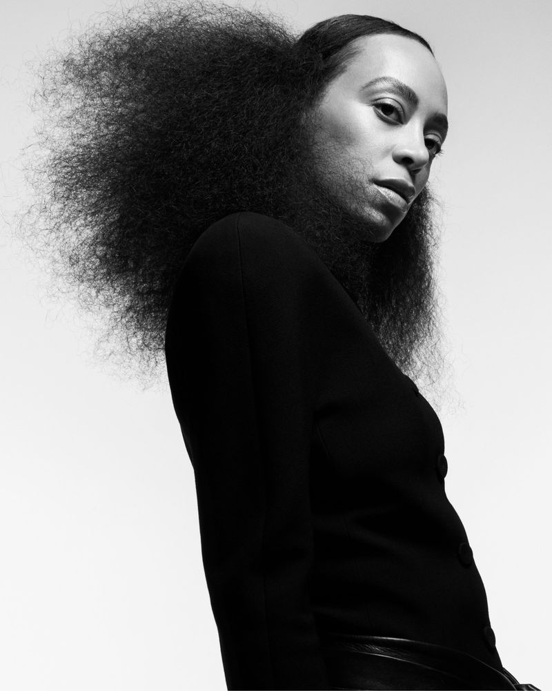 Black and white portrait of an adult female with large curly hair, looking at the camera, wearing a black long-sleeve shirt against a plain background.