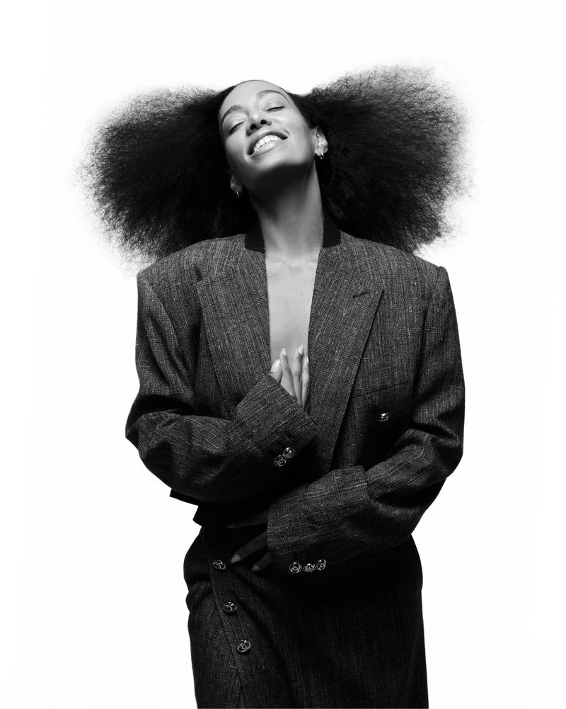 Black and white portrait of a smiling woman with large afro hair wearing a blazer and skirt with hands clasped. Isolated on white.