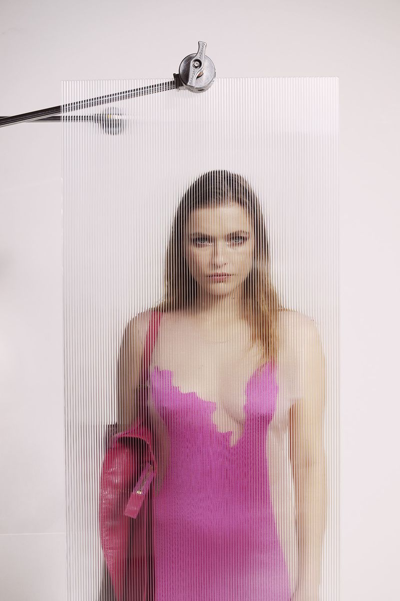 A young woman poses in a pink dress behind an optical grating filter, holding a red jacket in a photo studio with white background.