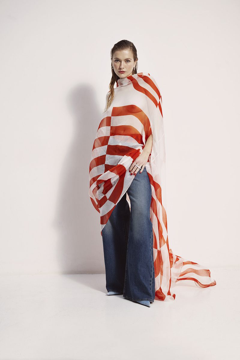 Full-length shot of a female model wearing wide-leg denim jeans and a red and white patterned poncho in a brightly lit studio.