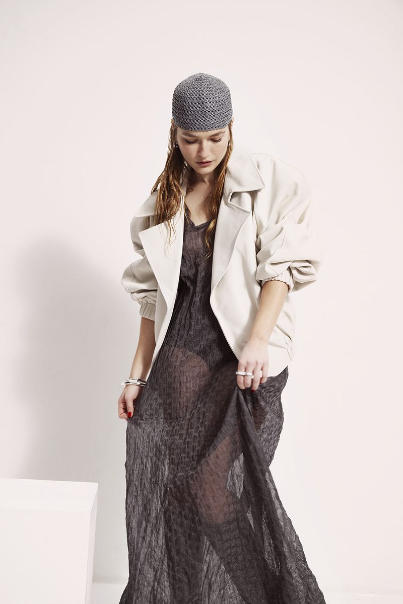 A fair-skinned adult woman wearing a gray beanie, sheer gray dress, and light jacket poses in a studio with a white background.