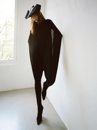 Adult woman in black jumpsuit and sculptural hat poses fashionably indoors against a white wall with a window letting in natural light.