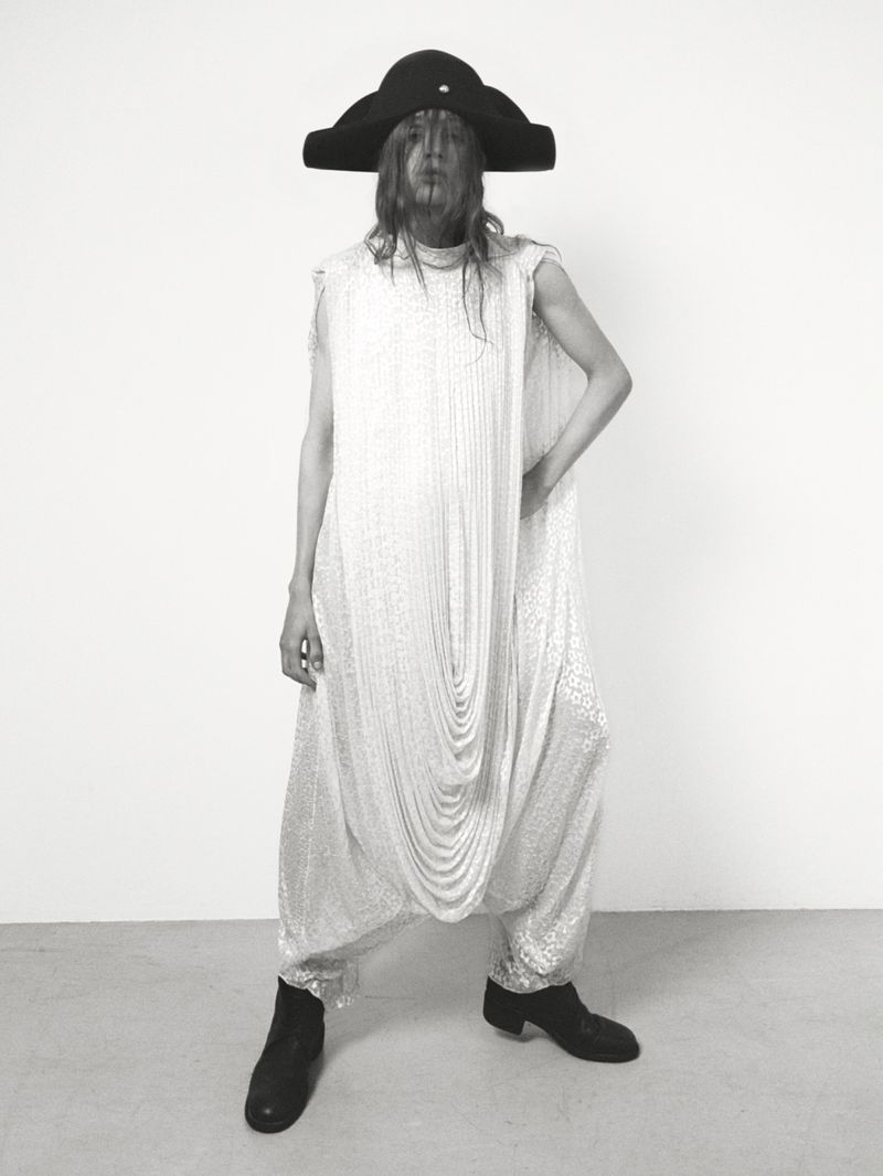 Person in a large hat and silver patterned jumpsuit stands in a photo studio, illuminated by neutral, diffused light.