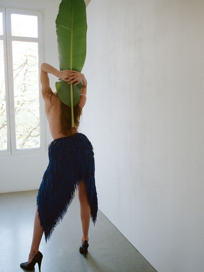 Back view of a partially nude woman in a fringed skirt holding a large green leaf, posing against a white wall and window.