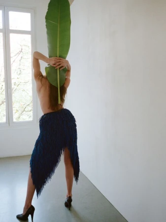 Back view of a partially nude woman in a fringed skirt holding a large green leaf, posing against a white wall and window.