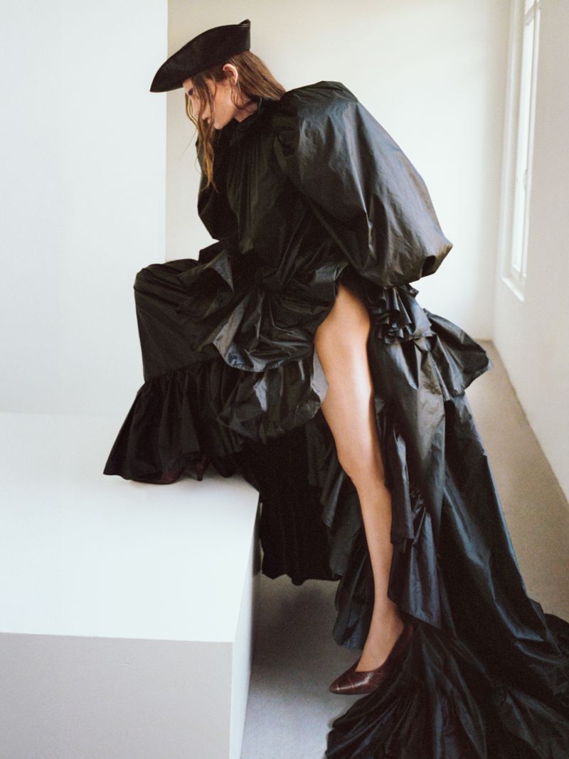 Fashion shot of an adult female in a voluminous black taffeta dress and black beret posing indoors on a white cube in a studio.