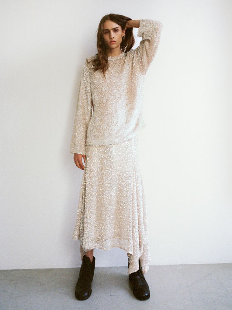 A slender model poses in a textured, long-sleeved top and asymmetrical skirt with black boots against a white studio backdrop.