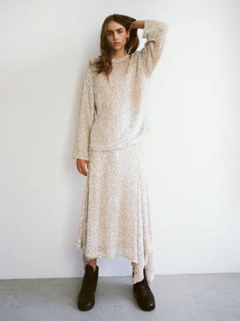 A slender model poses in a textured, long-sleeved top and asymmetrical skirt with black boots against a white studio backdrop.