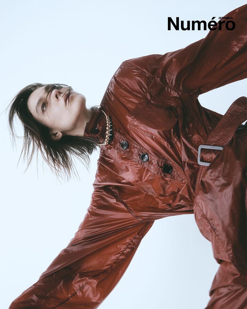 Fashion editorial shot: a slender adult female model with long brown hair poses indoors wearing a shiny rust-colored coat and necklace.