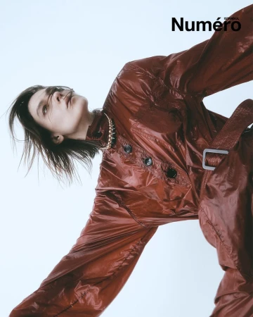 Fashion editorial shot: a slender adult female model with long brown hair poses indoors wearing a shiny rust-colored coat and necklace.