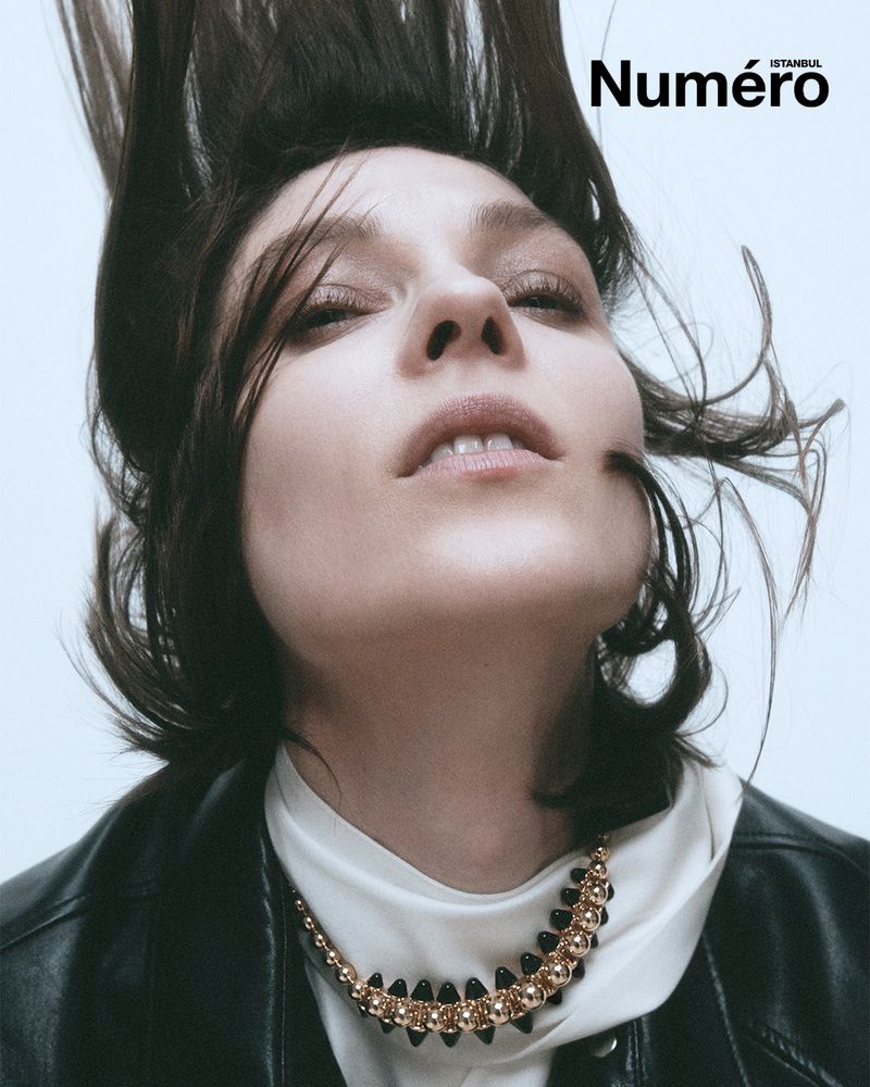 Fashion portrait of a woman with dark hair, wearing a necklace and black leather jacket, against a light background with logo.