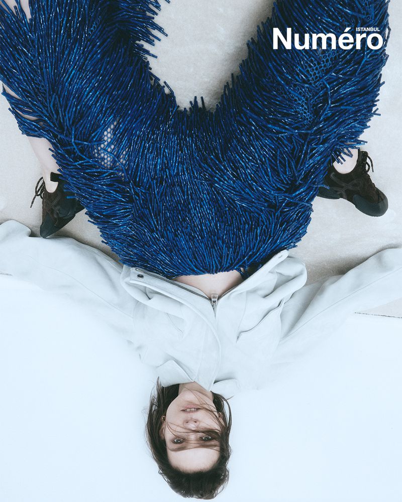 Model poses on a white floor wearing a hooded jacket and blue fringe pants in a fashion editorial shot for Numero Istanbul magazine.