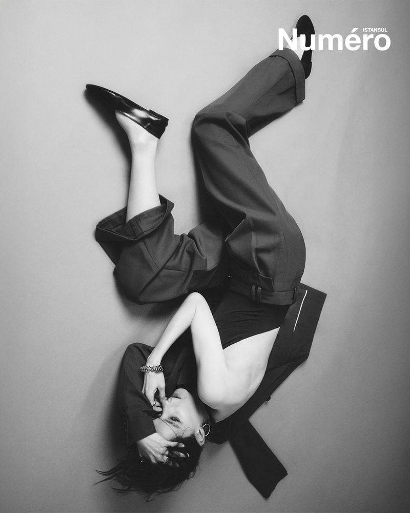 Black and white fashion editorial shot of a reclining woman in pants and heels against a grey background with the Numero magazine logo.