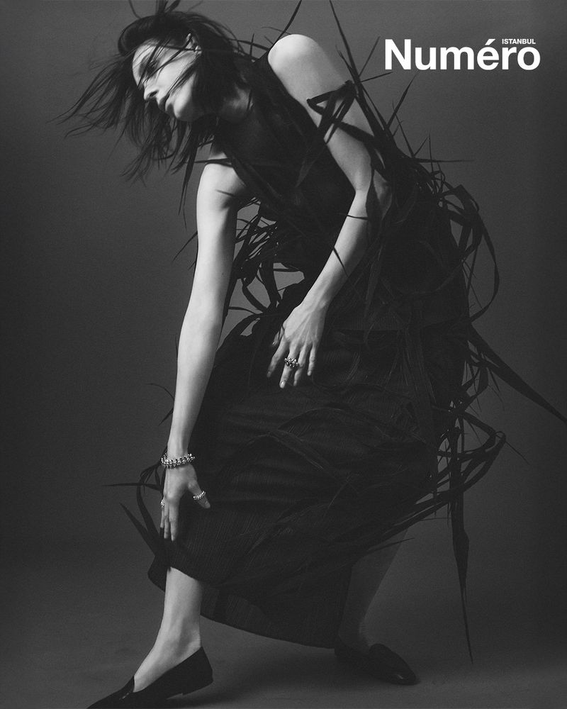 Fashion editorial shot in black and white featuring a female fashion model in a dark dress posing dramatically with wild-looking grass.