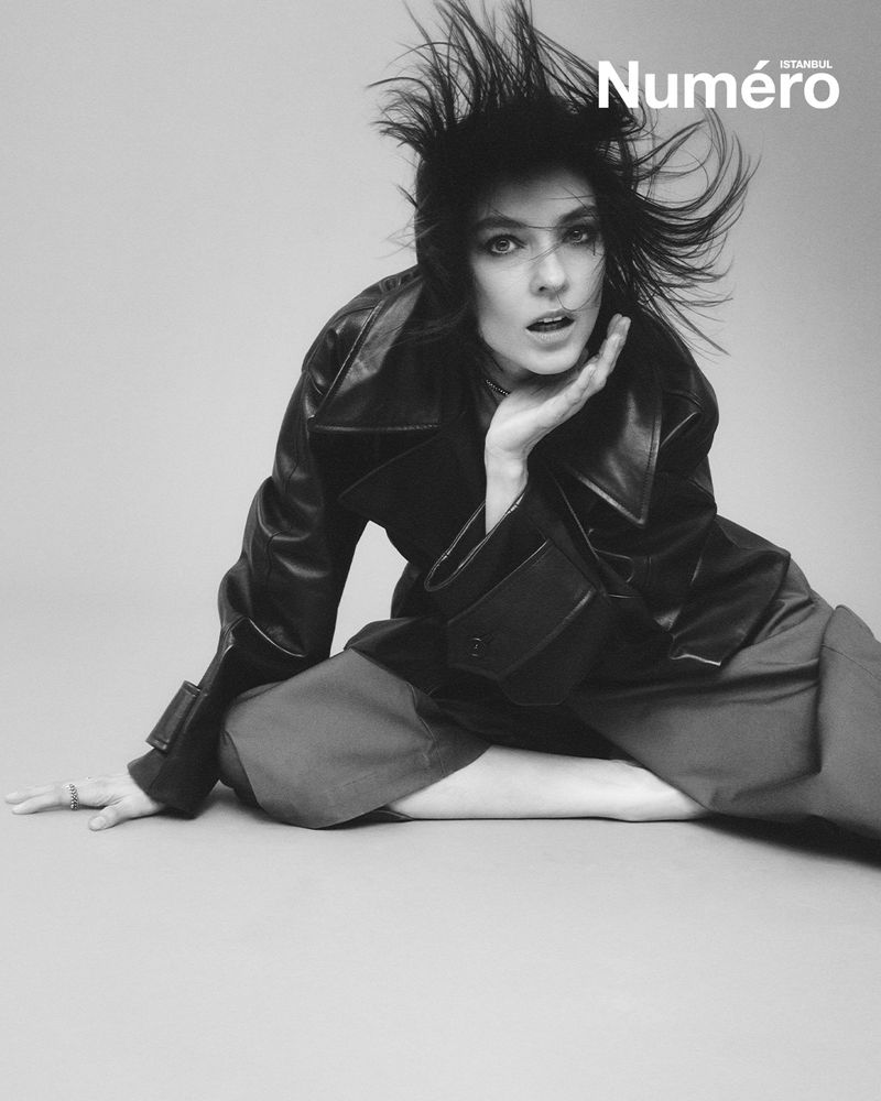 Black-and-white portrait of a woman with disheveled hair in a leather jacket and dark pants against a neutral backdrop.