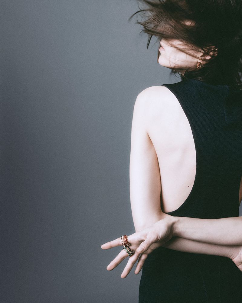 A woman in a black dress with her arms crossed behind her back, showcasing a detailed ring, against a gray background, indoor studio.