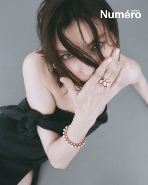 Close-up fashion portrait of a woman with dark hair wearing a black off-the-shoulder dress, gold jewelry, and posing against a grey background.