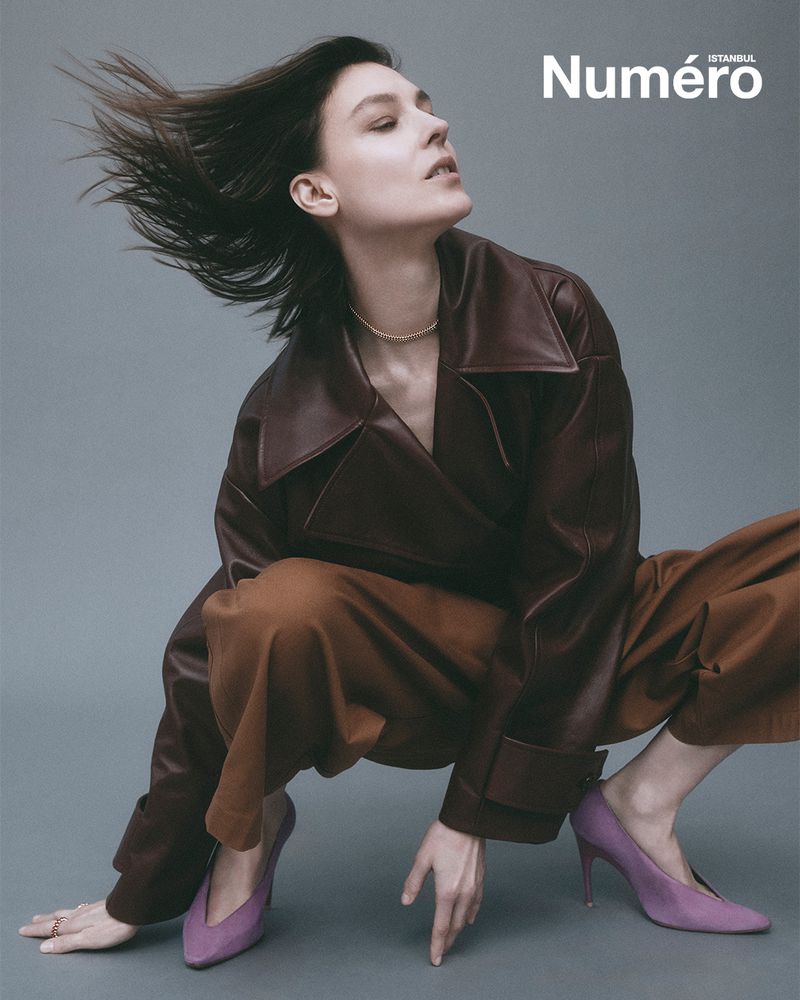 A young woman in a leather jacket and brown pants poses with wind-blown hair, wearing high heels, against a grey studio backdrop.