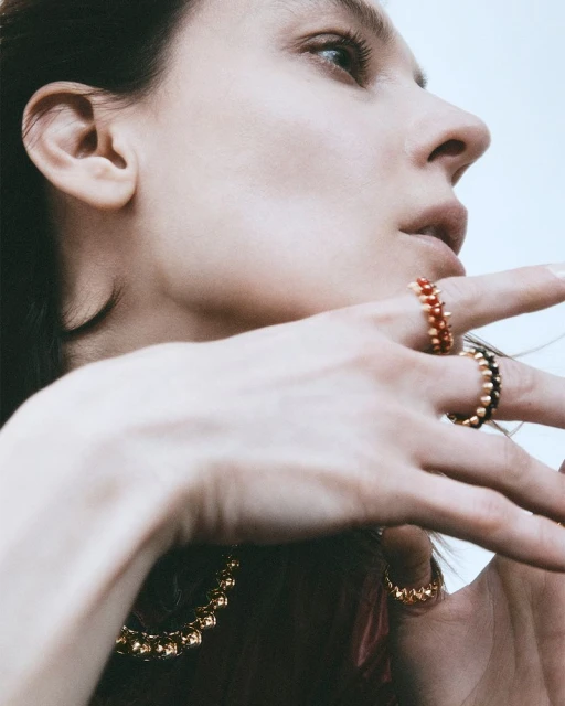Woman's profile wearing jewelry, including multiple gold beaded rings with red and black beads, and a gold beaded necklace.