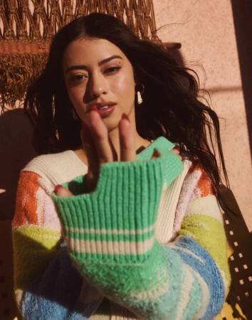 Portrait of an adult female model with dark hair wearing a colourful striped sweater posing indoors in front of a neutral wall.