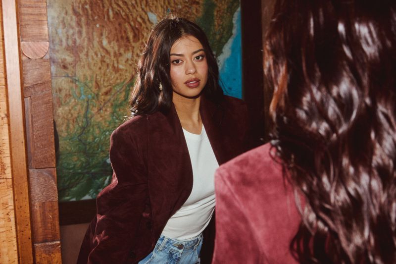Young woman in a burgundy blazer and white top poses indoors near a map and wood trim. Dark brown wavy hair frames her face.