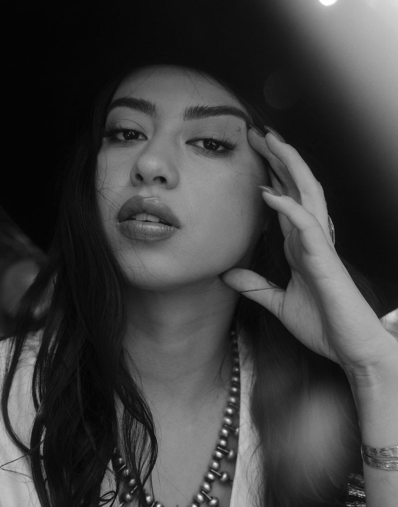 Monochrome portrait of an adult female model with long dark hair, wearing a beaded necklace and bracelets, posing in a dark studio.