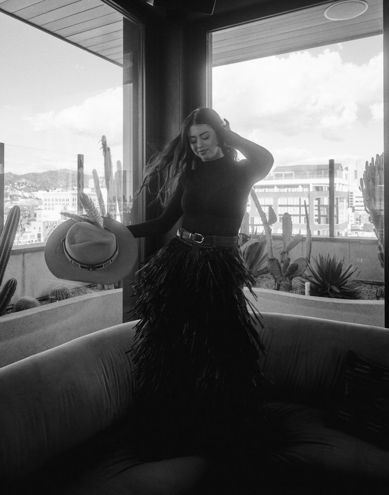 A woman poses on a couch near a window with cacti. She holds a hat and wears a fringed skirt and long-sleeved shirt in monochrome.