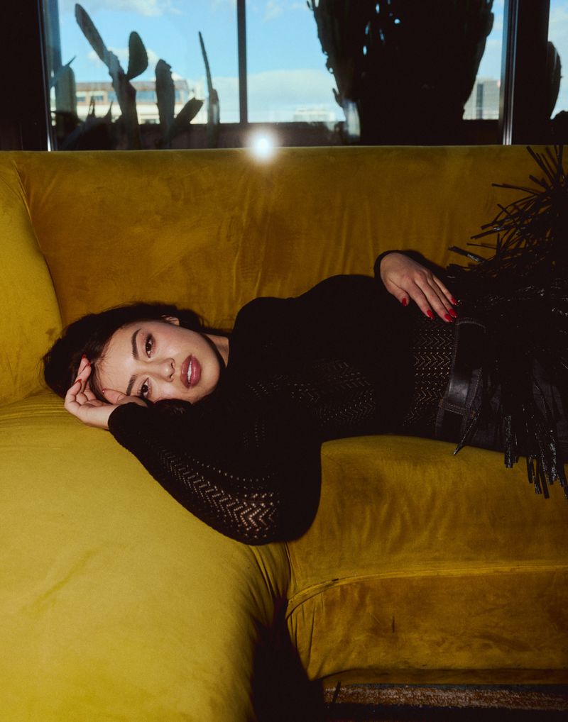 Brunette woman in black top lounges on a yellow velvet couch near a window with a city view in soft, warm lighting.