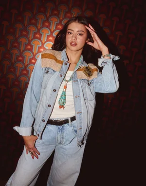 Amber Midthunder wearing a light wash denim jacket and jeans against a patterned orange wallpaper background.