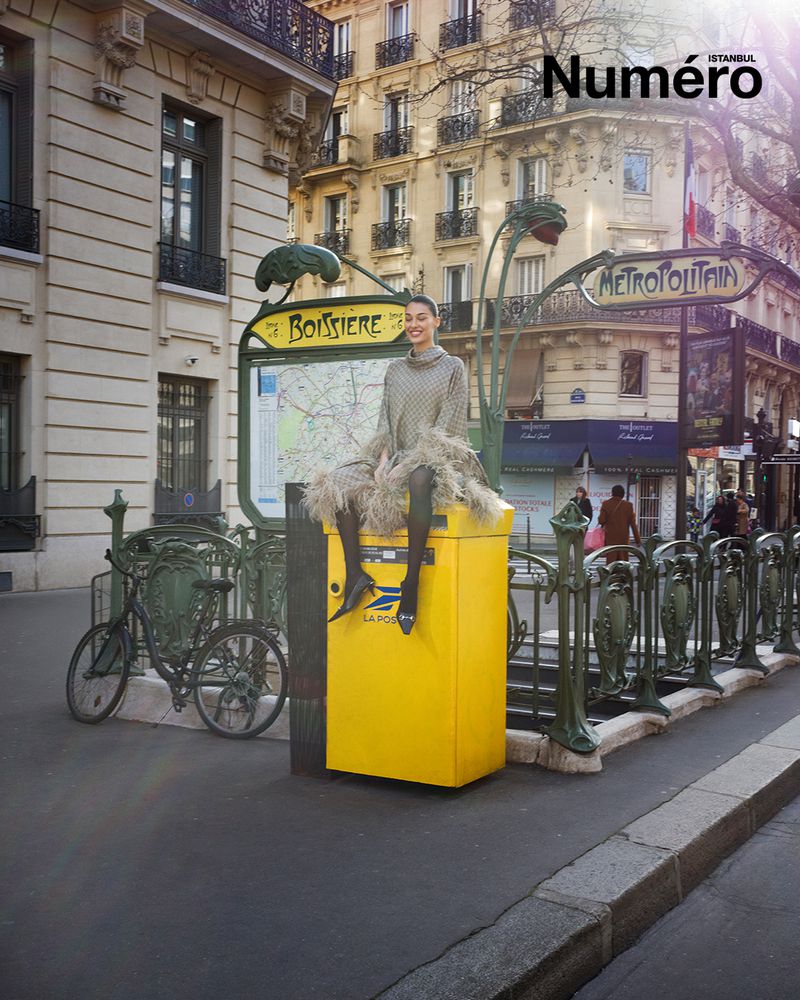 An adult woman in an ornate dress sits on a yellow La Poste mailbox near the Boissière Métro station in Paris on a sunny day.