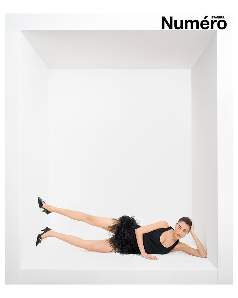 Fashion editorial shot of a woman in a black dress lying in a white box against a white background; fashion, style, pose, studio lighting.