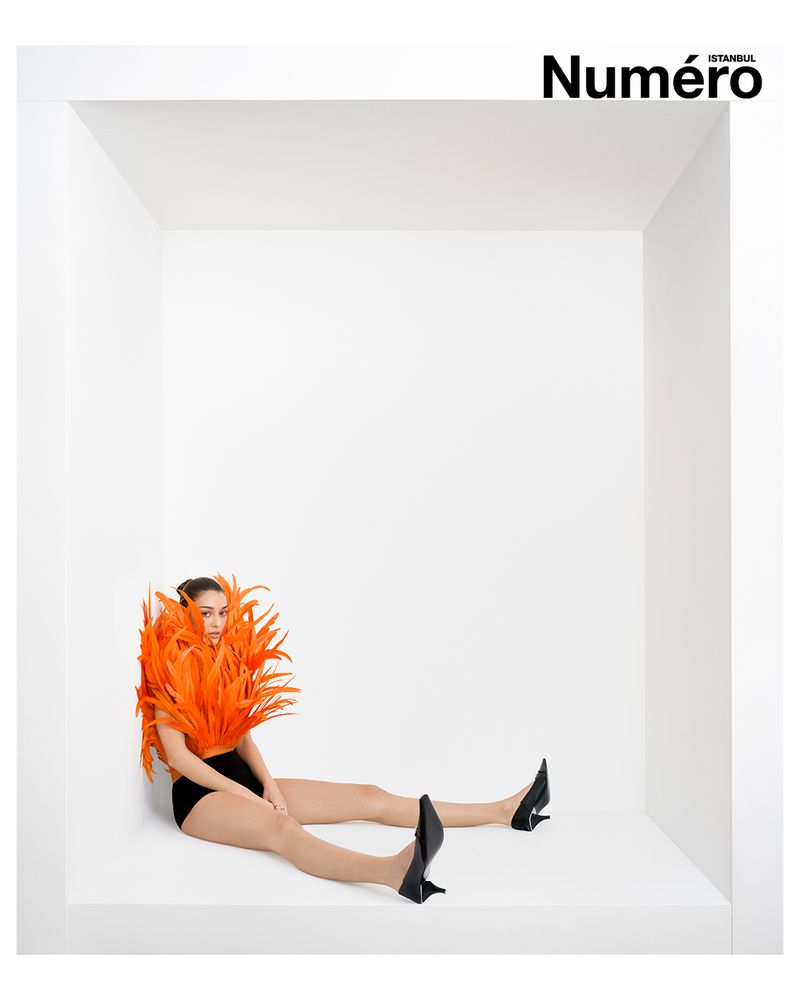 Editorial fashion shot of a woman in a black bodysuit and heels sitting in a white cube, adorned with orange feathers around her shoulders.