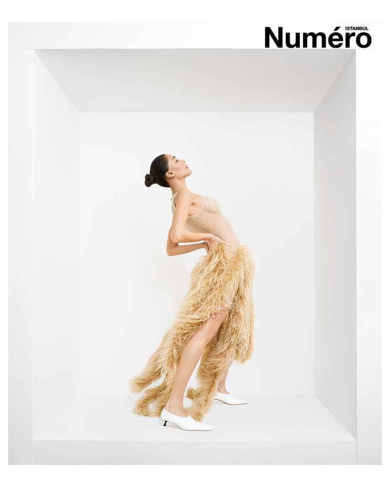 Fashion editorial shot of an adult female wearing a beige dress with feather details, white heels, posing in a white cube in studio.