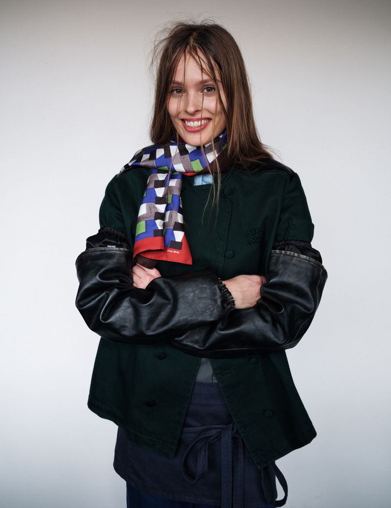 A smiling woman in a dark green jacket and patterned scarf stands against a white background, arms crossed in a studio setting.