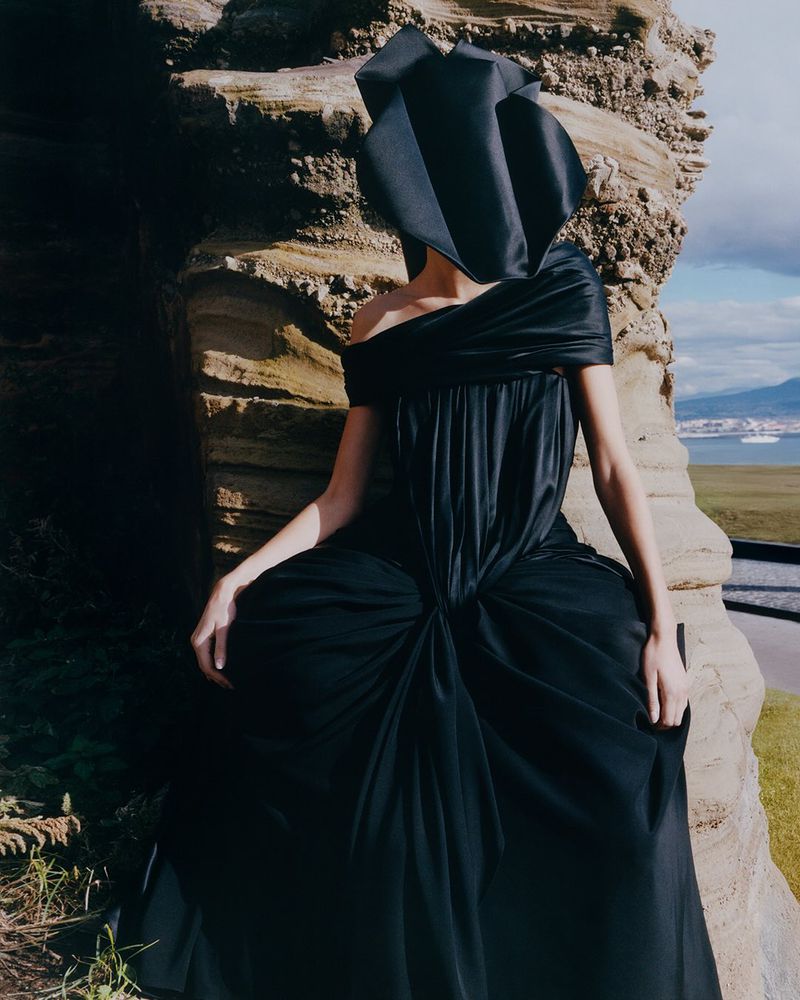 A person in a long, off-the-shoulder black gown and matching sculptural headpiece, stands near a natural rock formation with a sea view.