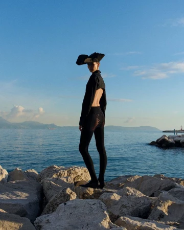Adult female fashion model poses on rocks wearing black cropped sweater, leggings, and large hat against a seaside background.