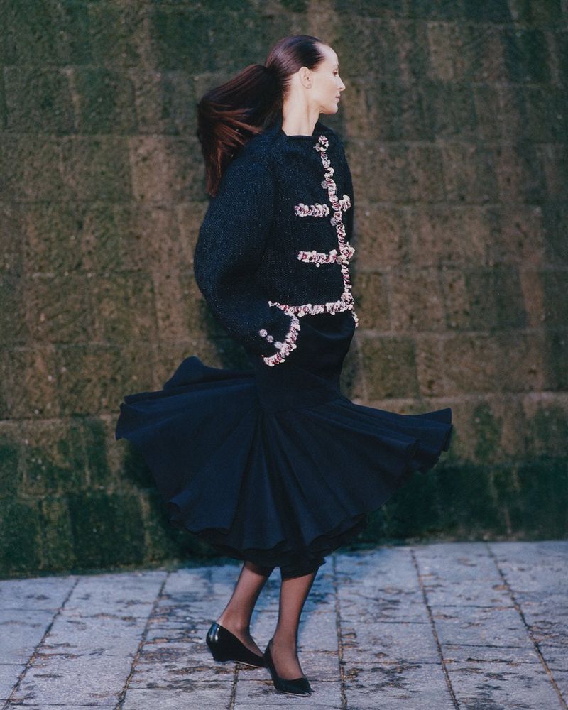 An adult female model poses outdoors in a dark, jeweled sweater and flowing skirt, complemented by black heels, in a fashion editorial shot.