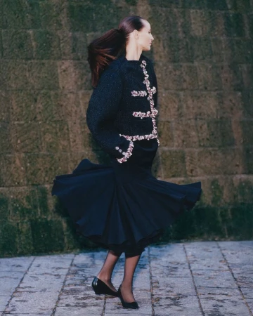 An adult female model poses outdoors in a dark, jeweled sweater and flowing skirt, complemented by black heels, in a fashion editorial shot.