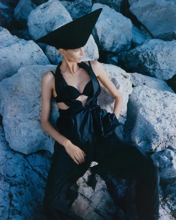 Fashion shot of a woman in a black outfit with a large black hat, lying on rocks in an outdoor setting, styled with dark tones.