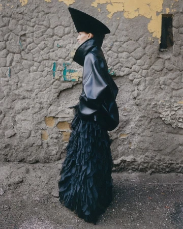 Fashionable woman in a sculptural black outfit with a dramatic hat poses against a textured grey wall, showcasing avant-garde style.