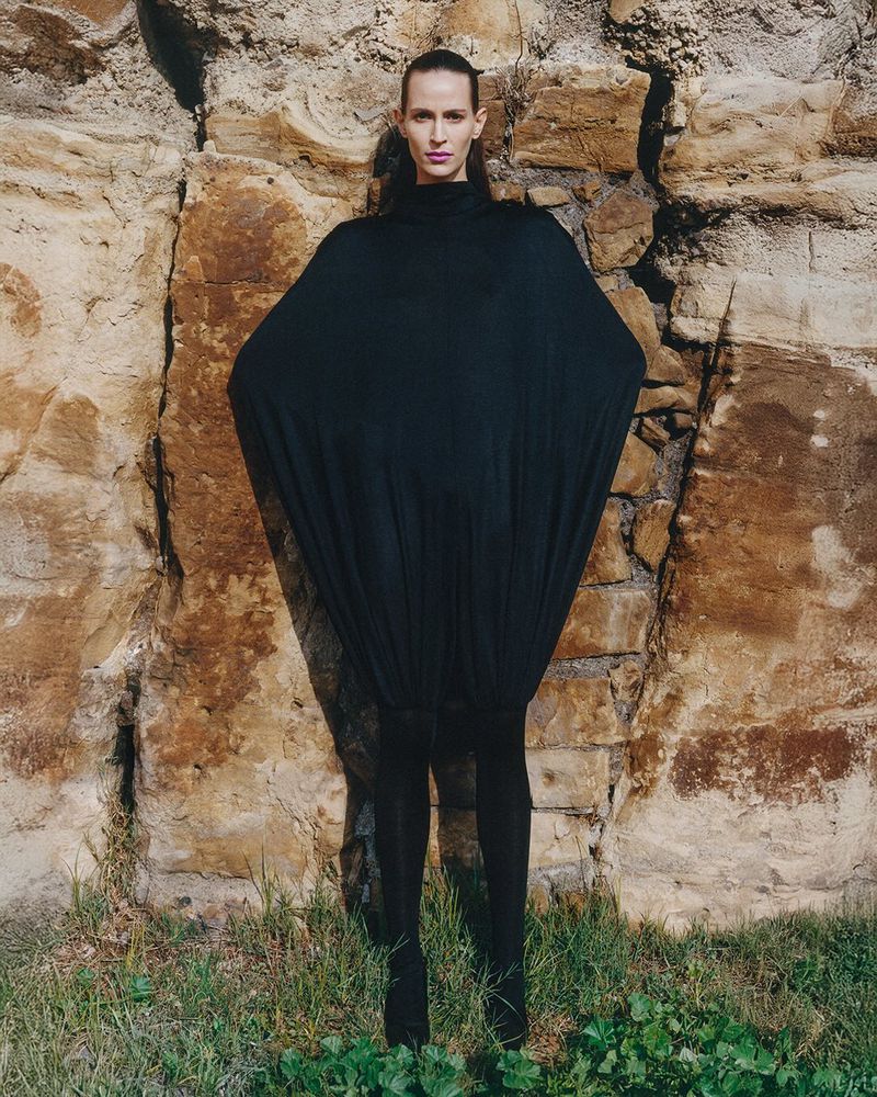 Adult female fashion model in oversized black draped dress, posing against a light brown stone wall and greenery on the ground.