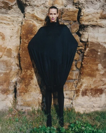 Adult female fashion model in oversized black draped dress, posing against a light brown stone wall and greenery on the ground.
