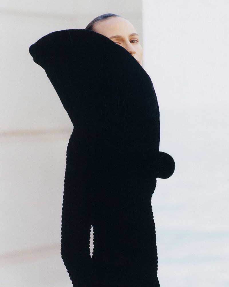 Woman with slicked-back hair poses in a black, long-sleeved, textured garment against a bright white, angular wall. Minimalist fashion.