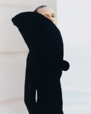 Woman with slicked-back hair poses in a black, long-sleeved, textured garment against a bright white, angular wall. Minimalist fashion.