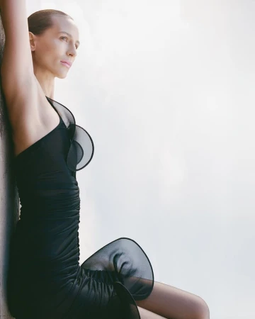 Adult woman in a one-shoulder black dress leans against a grey wall, gazing off-frame, captured in soft, natural lighting with neutral tones.