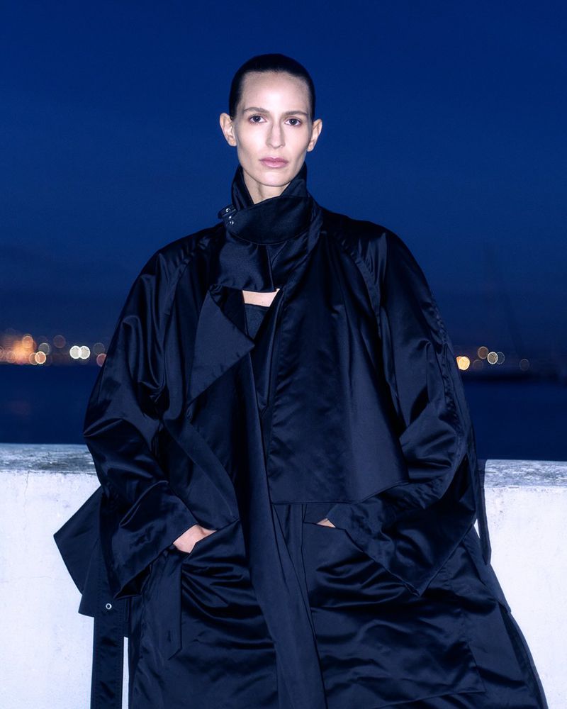 An adult female model wears a large black jacket in an outdoor evening setting with city lights and water in the background.
