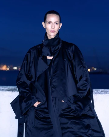An adult female model wears a large black jacket in an outdoor evening setting with city lights and water in the background.