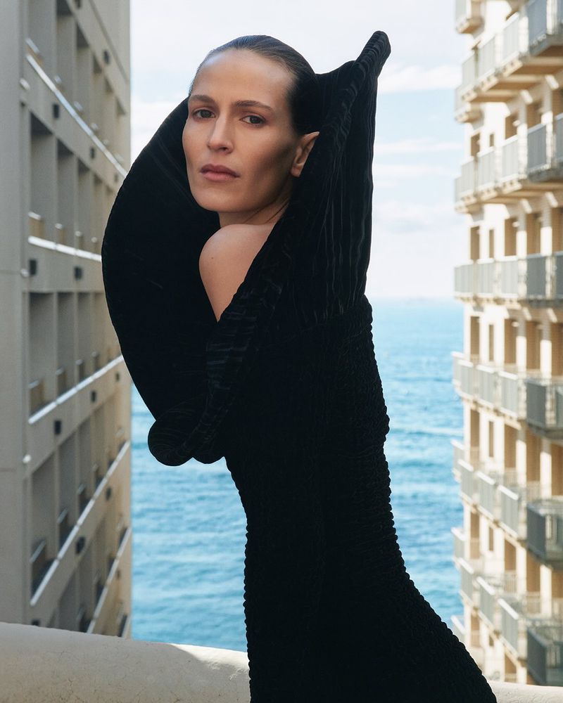 Elegant portrait of a woman in a black dress with a dramatic collar, posing outdoors against a backdrop of city buildings and ocean.