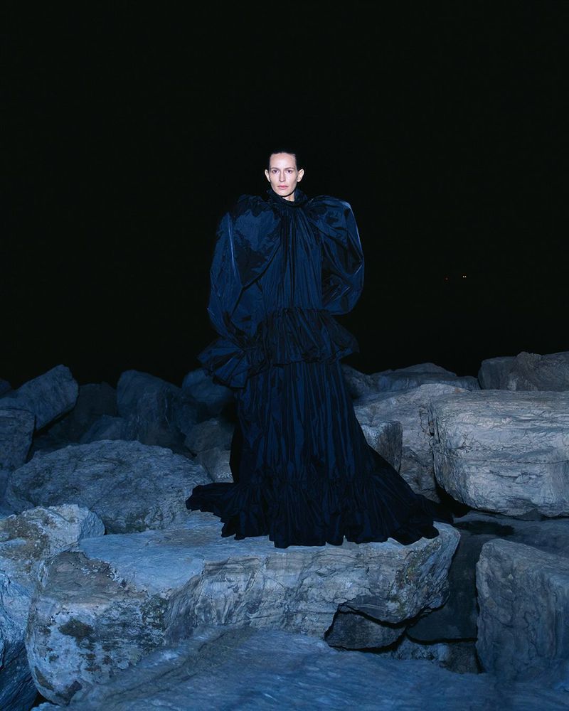 A single woman stands on rocks at night wearing a ruffled dark blue gown, under a spotlight in an outdoor environment.