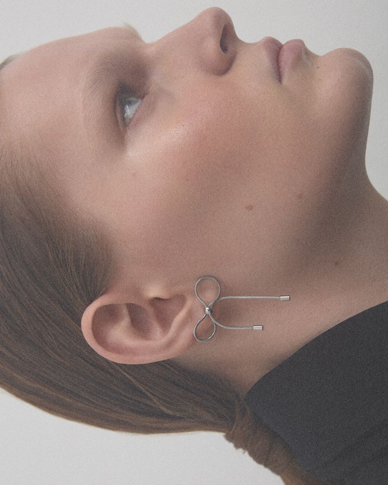 Elegant headshot of a young woman in a black shirt, wearing a creative metal earring shaped like a bow, against a neutral backdrop.